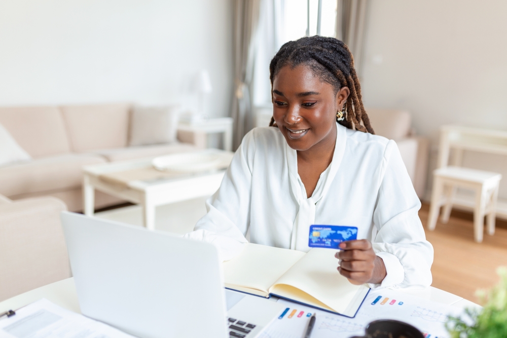 Beautiful smiling young African businesswoman holding a credit card and using a laptop in the office. 