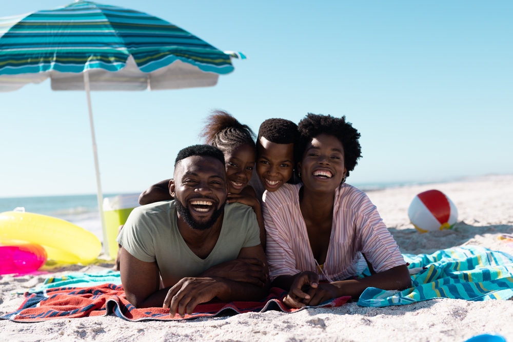 Cheerful african parents on a family friendly vactation destination with children lying on towels at beach under clear sky. Copy space, unaltered, family, together, parasol, picnic, nature, vacation, enjoyment, relaxing, summer.
