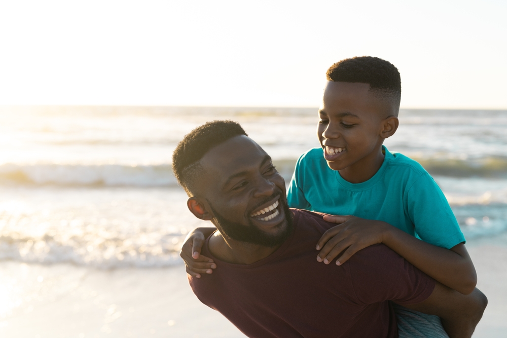 Cheerful african father piggybacking son against beautiful seascape and clear sky at sunset. Copy space, unaltered, family, together, childhood, nature, vacation, enjoyment and summer. 