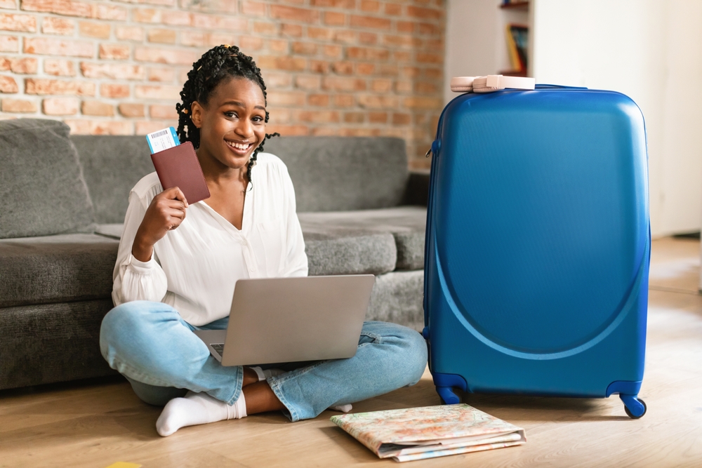 Positive african american woman showing passport and airplane ticket, using laptop to buy tourist vacation at internet travel agency, looking and smiling at camera while sitting neat suitcase 