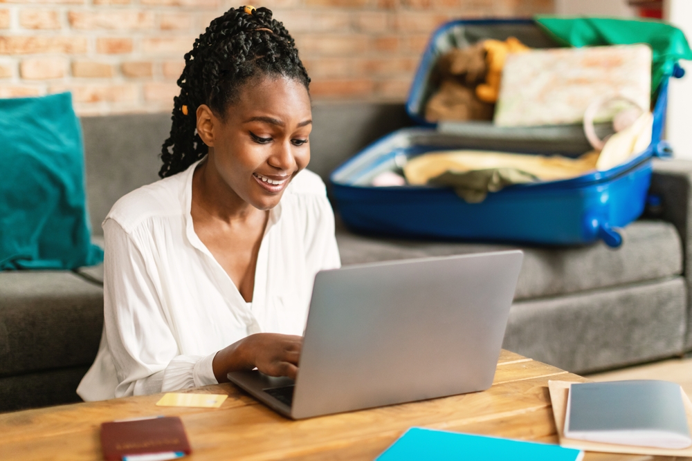 African woman planning vacation travel with laptop and searching for cheap summer flights from Nigeria , reading tourist blog online or booking hotel room on web, getting ready for abroad journey at home 