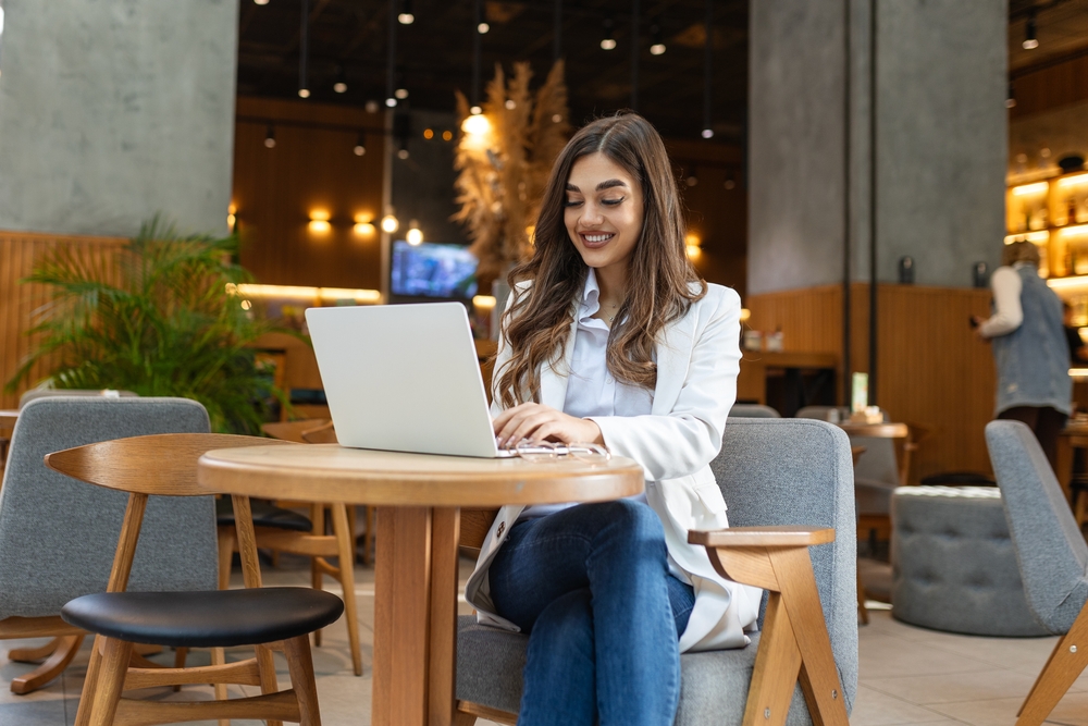 Young woman, digital nomad visa lady working remotely from a cafe, drinking coffee and using laptop, smiling.