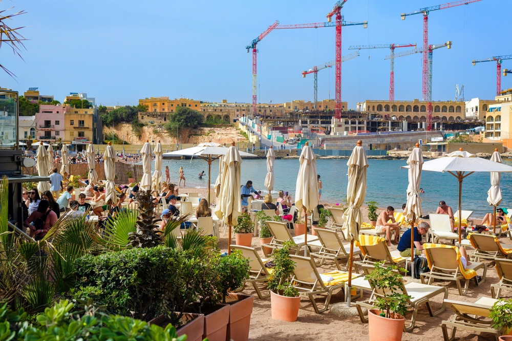 St. Julian's, Malta- 04.15.2025: A busy beach in Malta with sunbathers, umbrellas, and loungers set against the backdrop of active construction and cranes — a contrast of tourism and urban growth.
